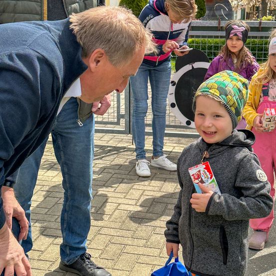Ein Mann und ein kleiner Junge stehen vor einem Metallzaun. Der Junge hält eine blaue Tasche und lächelt. Hinter ihnen schauen eine Frau und ein Mädchen auf ihre Handys. Das Mädchen trägt eine Mütze.