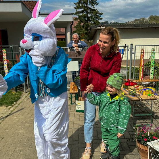 Eine Frau und ein Kind stehen auf einem gepflasterten Bereich mit einer Person in einem Hasenkostüm, einem Tisch mit einer Topfpflanze und einem Mann hinter einem Zaun.
