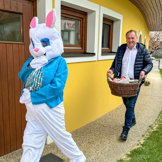 Person in Osterhasenkostüm mit einem Korb voller Ostereier vor einem gelben Gebäude, hinter dem ein Mann geht.