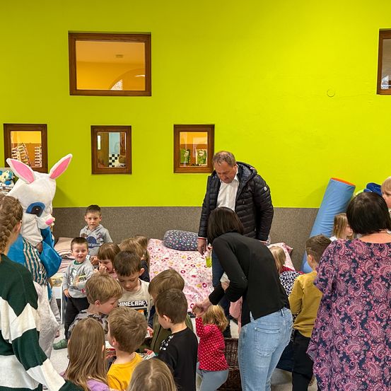 Eine Gruppe von Kindern und Erwachsenen versammelt sich um eine Person in einer Bunny-Maskottchen-Verkleidung, eine Person hält einen Korb und eine Person einen Kissen. Es hängen Bilderrahmen an einer grünen Wand.