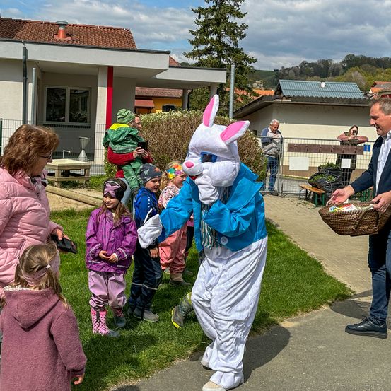 Ein Mann in einem Osterhasenkostüm verteilt Ostereier an eine Gruppe von Kindern und ihren Eltern in einem Garten.