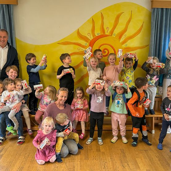 Eine Gruppe von Kindern und Erwachsenen in einem Raum mit einer Sonne an der Wand. Die Kinder halten Boxen mit Snacks hoch. Einige stehen und andere sitzen. Ein Maskottchen steht ganz rechts.