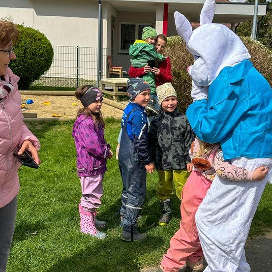 Eine Frau mit einem Tablet spricht zu einer Gruppe von Kindern, eines in einem Osterhasenkostüm, auf einer Rasenfläche.