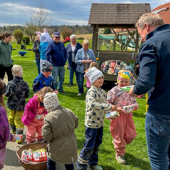 Kinder bekommen Ostereier von einem Mann vor einem Osterhasen, mit Erwachsenen und einem Spielplatz im Hintergrund.