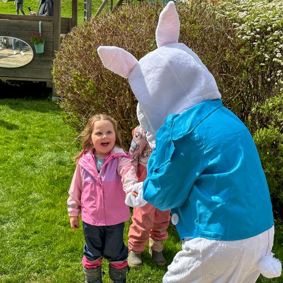 Ein kleines Mädchen mit einer rosa Jacke lacht und hält die Hand eines Erwachsenen, der als Hase verkleidet ist, auf einem grünen Rasen.