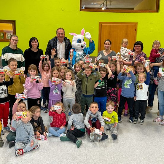 Eine Gruppe von Kindern und Erwachsenen posiert für ein Foto in einem Raum mit gelben Wänden. Die Kinder halten Schachteln mit Süßigkeiten in der Hand, und einige von ihnen tragen Turnschuhe. Ein Maskottchen mit Hasenohren steht hinter ihnen, und eine Person in einem Anzug hält ein Kind.