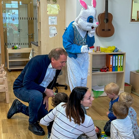 Ein Mann in Anzug und eine Frau in einem gestreiften Shirt sitzen mit drei Kindern auf dem Boden. Ein Erwachsener als Osterhase steht hinter ihnen.