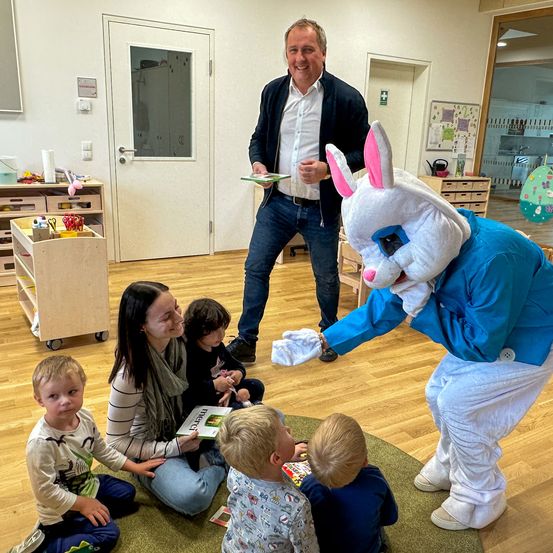 Eine in einen Hasenkostüm verkleidete Person überreicht einer Frau und Kindern, die auf dem Boden sitzen, ein Buch.