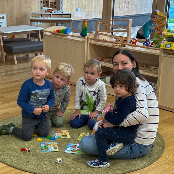 Eine Frau sitzt mit drei Jungen und einem kleinen Mädchen auf dem Boden und spielt mit Spielzeug. Die Frau lächelt.