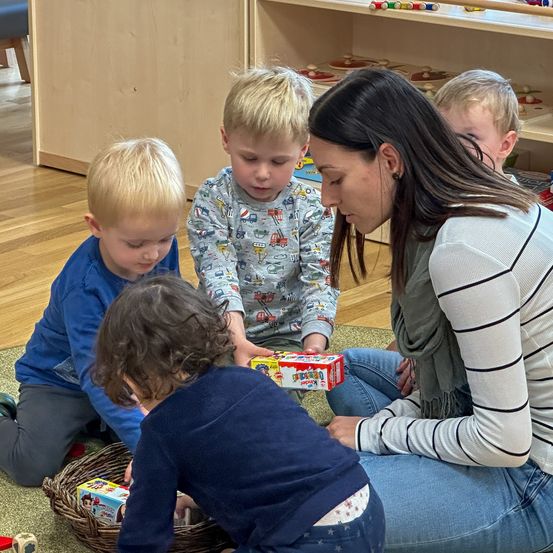Vier Kinder spielen mit Spielzeug, während eine Frau sie beobachtet. Die Frau sitzt auf dem Boden mit einem Korb voller Spielzeug vor sich.