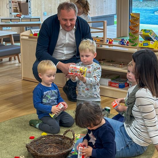 Ein Mann sitzt mit vier Kindern auf einem Teppich. Sie halten alle Spielzeug in den Händen. Der Mann trägt eine dunkelblaue Jacke und ein weißes Hemd. Die Kinder tragen langärmelige Oberteile.