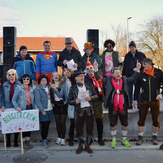 Eine Gruppe von Menschen in Kostümen und Hüten steht auf einer Straße, hält Schilder und singt, mit Lautsprechern und einem Gebäude im Hintergrund.