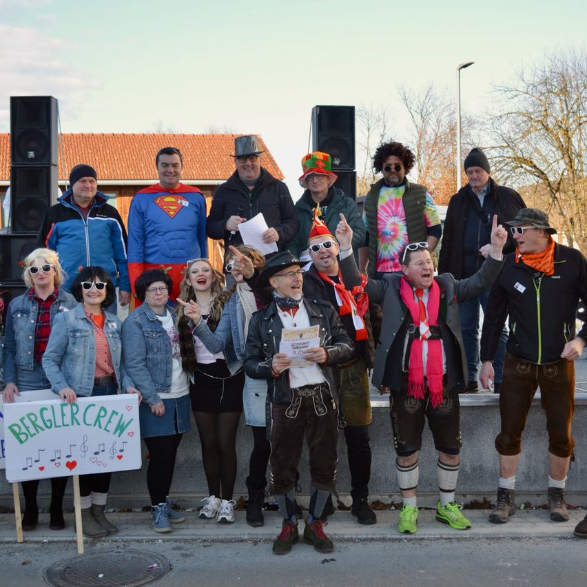 Eine Gruppe von Menschen in Kostümen und Hüten steht auf einer Straße, hält Schilder und singt, mit Lautsprechern und einem Gebäude im Hintergrund.
