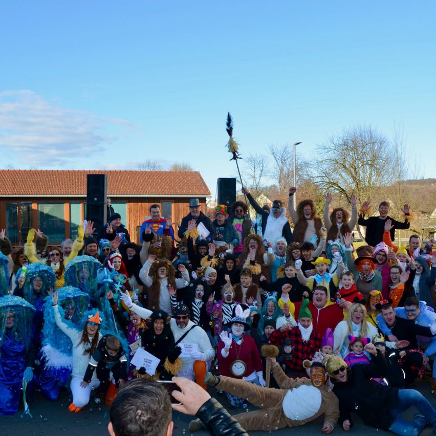 Eine große Gruppe von Menschen in verschiedenen Kostümen versammelt sich im Freien. Viele winken und posieren für ein Foto. Ein Gebäude mit einem braunen Dach ist im Hintergrund.