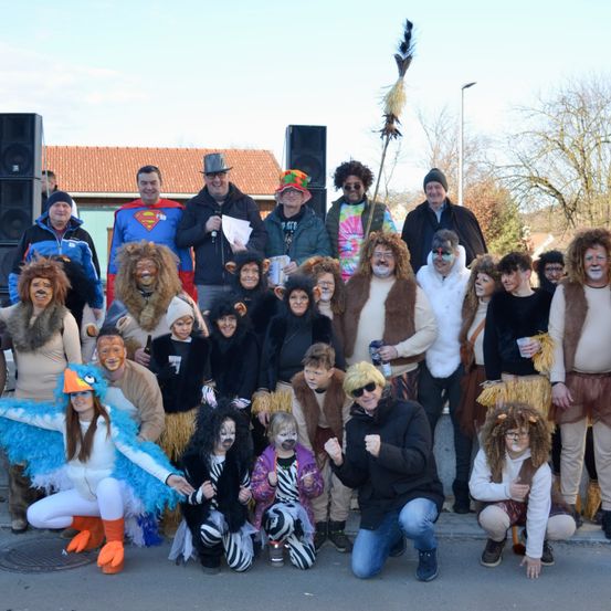 Eine Gruppe von Menschen posiert für ein Foto draußen, einige in Tierkostümen und andere in Superheldenkostümen. Im Hintergrund befinden sich Lautsprecher und ein Mann mit einem Mikrofon.