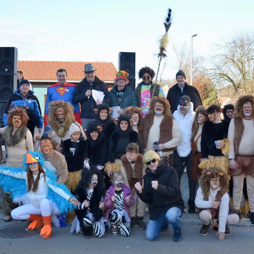 Eine Gruppe von Menschen posiert für ein Foto draußen, einige in Tierkostümen und andere in Superheldenkostümen. Im Hintergrund befinden sich Lautsprecher und ein Mann mit einem Mikrofon.