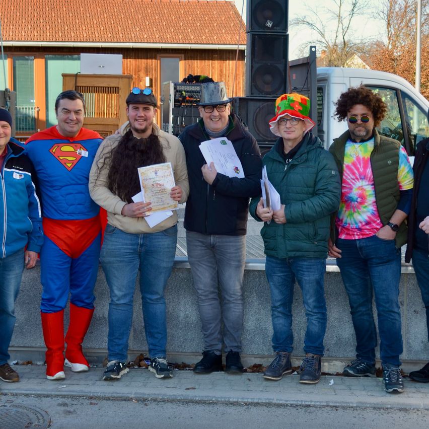 Eine Gruppe von Menschen in Kostümen posiert für ein Foto auf einem Bürgersteig und hält Urkunden in der Hand. Dahinter steht ein Gebäude, ein Van und Bäume.