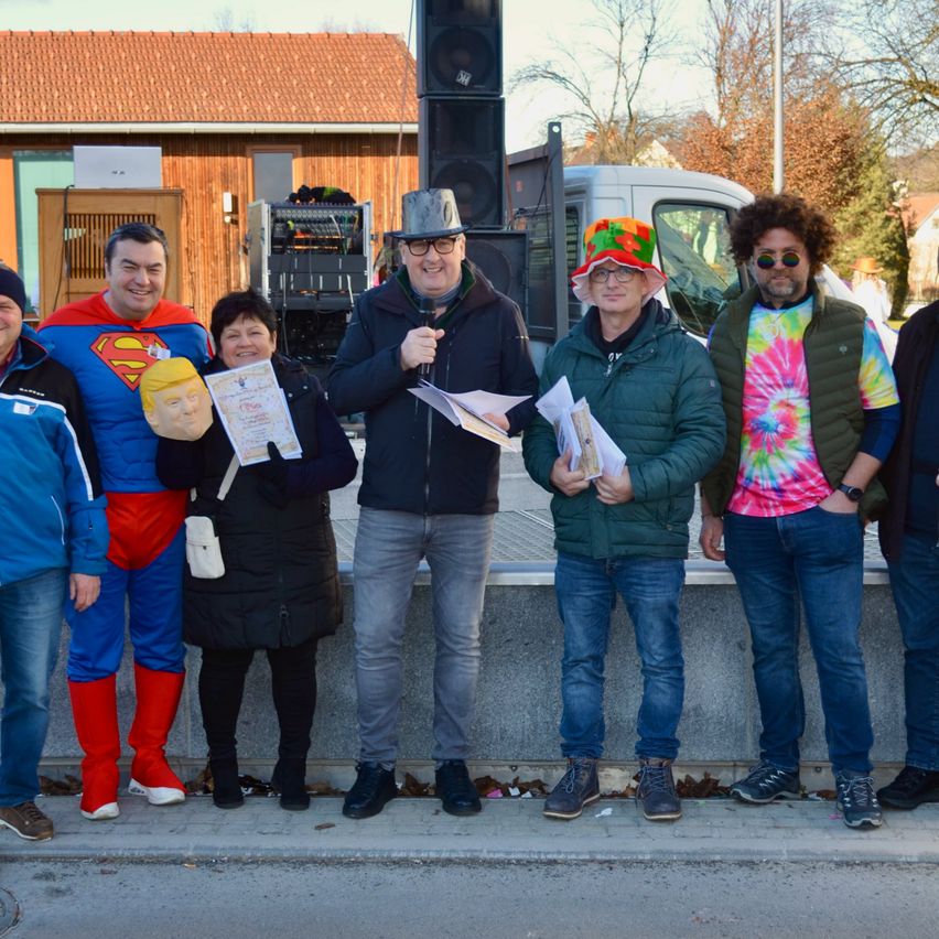 Eine Gruppe von Menschen in verschiedenen Kostümen, einschließlich eines Superman-Kostüms, steht zusammen vor einem Gebäude mit einem Lkw und Lautsprechern im Hintergrund.