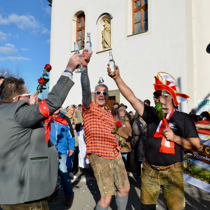 Eine Gruppe von Männern in traditioneller Kleidung feiert, hält Flaschen und Rosen. Einer trägt einen Hut, und sie stehen vor einem weißen Gebäude mit Fenstern und einer Statue.