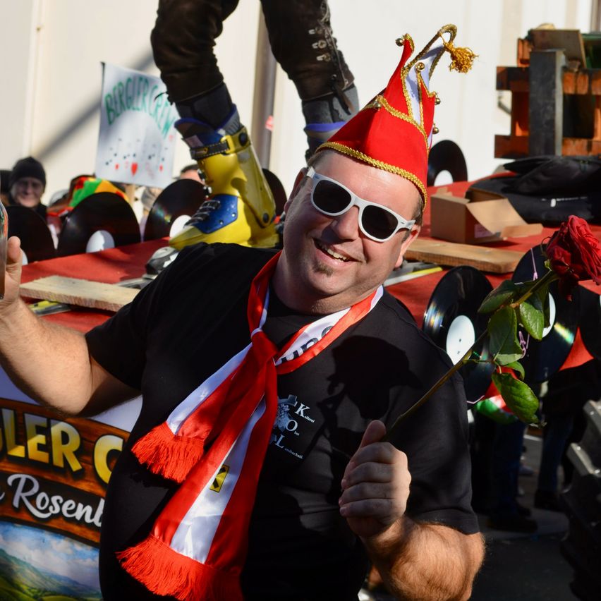 Ein Mann mit rotem Hut, Sonnenbrille und rot-weißem Schal hält eine Rose vor einem Umzugswagen.