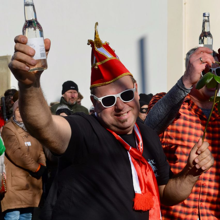 Ein Mann in einem roten Hut und einer Sonnenbrille hebt eine Flasche hoch, während ein anderer Mann hinter ihm eine Rose hält. Sie sind von anderen in einer festlichen Umgebung umgeben.
