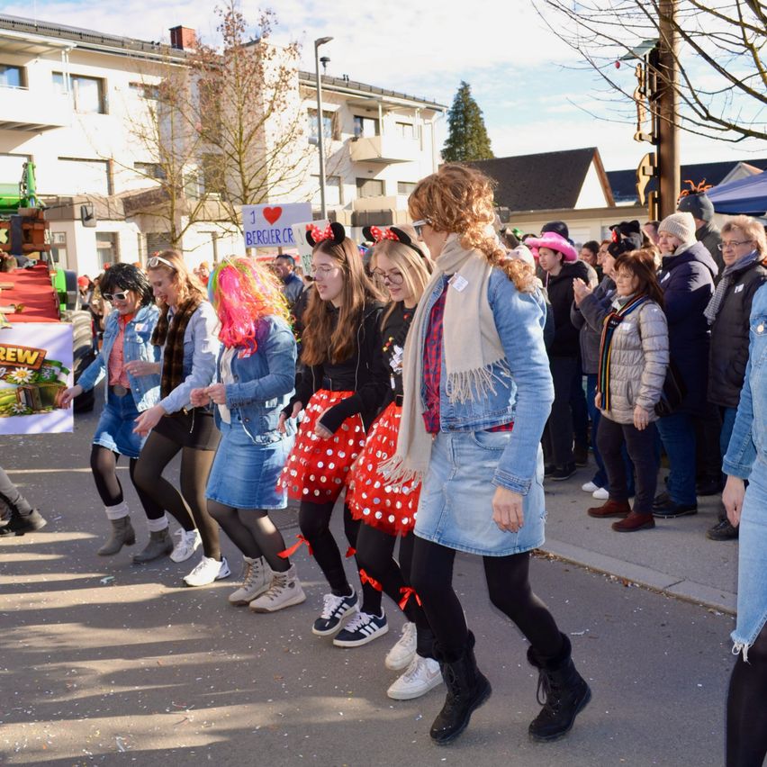 Eine Gruppe von Frauen in Jeans und gepunkteten Röcken tanzt auf der Straße, umgeben von Schaulustigen während einer Parade. Die Frauen tragen Stiefel und Schals, und ein Banner mit dem Wort 'CREW' ist im Hintergrund sichtbar.