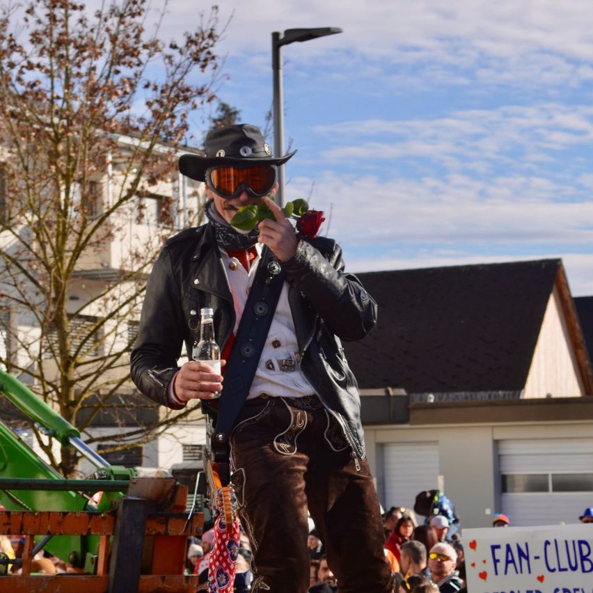Ein Mann in Cowboy-Kleidung steht auf einem Anhänger, hält eine Flasche und eine Rose und hat eine Menge und Gebäude im Hintergrund.