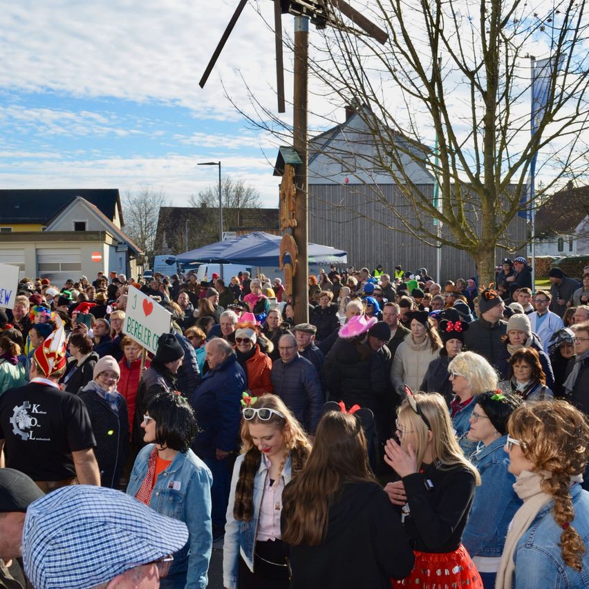 Eine große Menschenmenge versammelt sich im Freien unter blauem Himmel, viele tragen Hüte und Brillen. Einige halten Schilder hoch, und ein Holzpfosten steht im Vordergrund. Gebäude sind im Hintergrund zu sehen.