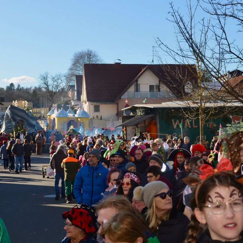 Eine große Menschenmenge in festlichen Kostümen versammelt sich auf der Straße, umgeben von Häusern und Gebäuden. Im Hintergrund ist ein Gebäude mit dem Wort 'WAL' und eine große Schuhskulptur zu sehen.