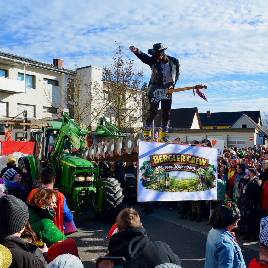 Ein Musiker steht auf einem Traktor bei einem Umzug. Die Menschen um ihn herum sind in festlichen Kostümen gekleidet. Ein von jemandem in der Menge gehaltenes Banner trägt den Schriftzug 'Bergler Crew vom Rosenberg'. Im Hintergrund sind Gebäude zu sehen.
