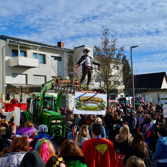 Ein Mann in Cowboyhut und Stiefeln steht auf einer Plattform über einer Menschenmenge. Er hält ein Schild mit den Worten Berg & Crew. Ein Traktor ist hinter ihm geparkt. Die Menschen sind in Kostümen gekleidet und stehen um ihn herum.