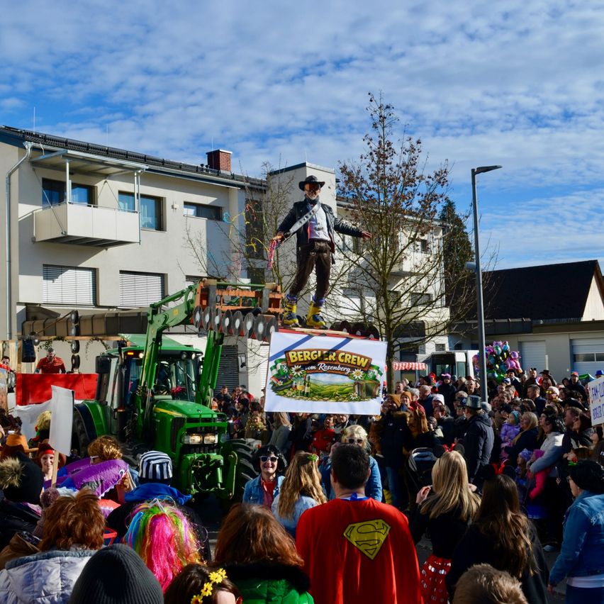 Ein Mann in Cowboyhut und Stiefeln steht auf einer Plattform über einer Menschenmenge. Er hält ein Schild mit den Worten Berg & Crew. Ein Traktor ist hinter ihm geparkt. Die Menschen sind in Kostümen gekleidet und stehen um ihn herum.