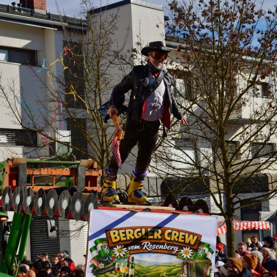 Ein Mann, gekleidet in einer Lederjacke und gelben Stiefeln, steht auf einer erhöhten Plattform mit einem Schild, das 'Bergler Crew vom Rosenberg' vor einem Gebäude mit vielen Fenstern steht. Dahinter ist ein blattloser Baum. Viele Menschen beobachten ihn.
