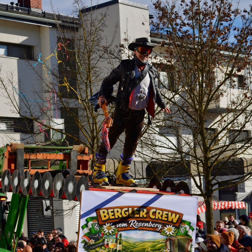 Ein Mann, gekleidet in einer Lederjacke und gelben Stiefeln, steht auf einer erhöhten Plattform mit einem Schild, das 'Bergler Crew vom Rosenberg' vor einem Gebäude mit vielen Fenstern steht. Dahinter ist ein blattloser Baum. Viele Menschen beobachten ihn.