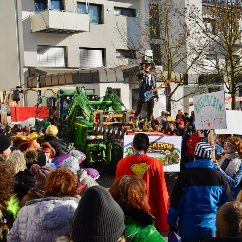 Eine Menschenmenge versammelt sich auf einer Straße und beobachtet einen Mann, der auf einem Traktor eine Gitarre spielt. Der Mann trägt eine Sonnenbrille und eine Lederjacke. Die Menge ist in Winterkleidung gekleidet. Ein Schild steht 'Biegler Crew'. Hinter dem Traktor befindet sich ein Gebäude mit vielen Fenstern.