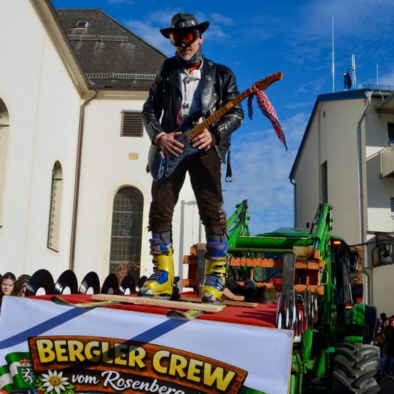 Ein Mann in einer Lederjacke, mit Schutzbrille und Stiefeln steht auf einem Traktor und spielt Gitarre. Dahinter befindet sich ein Gebäude mit Schornstein.