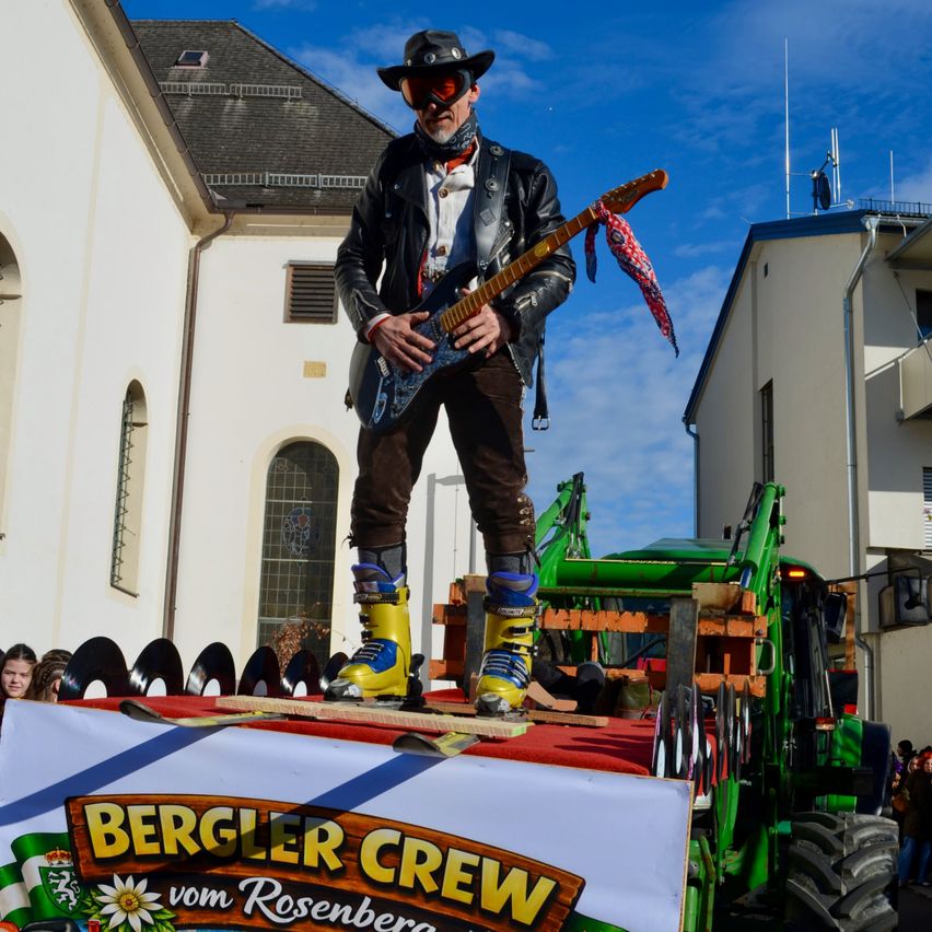 Ein Mann in einer Lederjacke, mit Schutzbrille und Stiefeln steht auf einem Traktor und spielt Gitarre. Dahinter befindet sich ein Gebäude mit Schornstein.