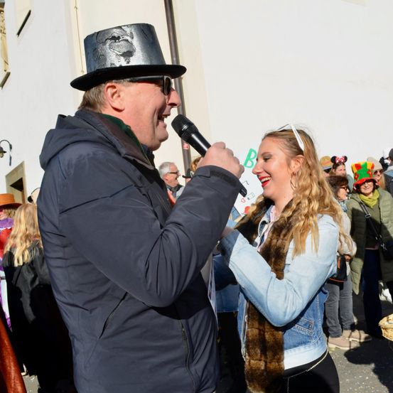 Ein Mann in einem schwarzen Hut mit einem Mikrofon spricht zu einer lächelnden Frau in einer Jeansjacke, umgeben von einer Menschenmenge in festlichen Hüten und Kostümen.