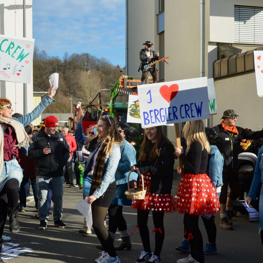 Eine Gruppe von Menschen marschiert in einem Umzug und hält Schilder und Körbe. Ein Schild steht 'J I BERGLER CREW'. Eine Person spielt Gitarre auf einem Wagen im Hintergrund. Bäume und Gebäude sind in der Ferne zu sehen.