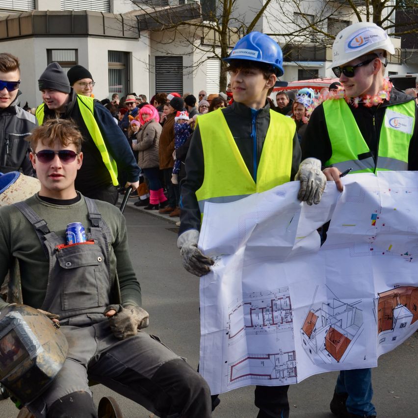 Drei junge Männer in Bauarbeiterkleidung gehen durch eine Menschenmenge. Einer hält eine Blaupause, während ein anderer ein Stück Papier in der Hand hat. Der dritte Mann sitzt auf einem Stuhl mit einem Schweißerhelm.
