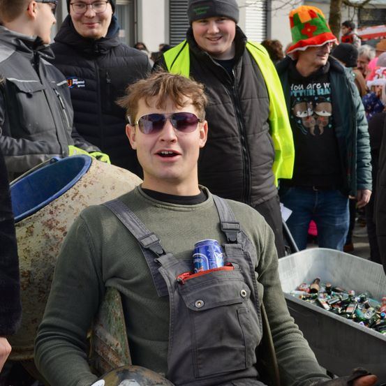 Eine Gruppe von Menschen ist in einer Außenumgebung versammelt. Ein Mann in einer Latzhose hält eine Dose in seiner Tasche. Ein anderer Mann in einer reflektierenden Weste und Mütze lächelt. Im Hintergrund trägt ein anderer Mann einen bunten Hut.