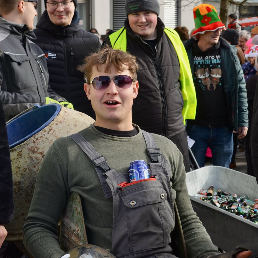 Eine Gruppe von Menschen ist in einer Außenumgebung versammelt. Ein Mann in einer Latzhose hält eine Dose in seiner Tasche. Ein anderer Mann in einer reflektierenden Weste und Mütze lächelt. Im Hintergrund trägt ein anderer Mann einen bunten Hut.