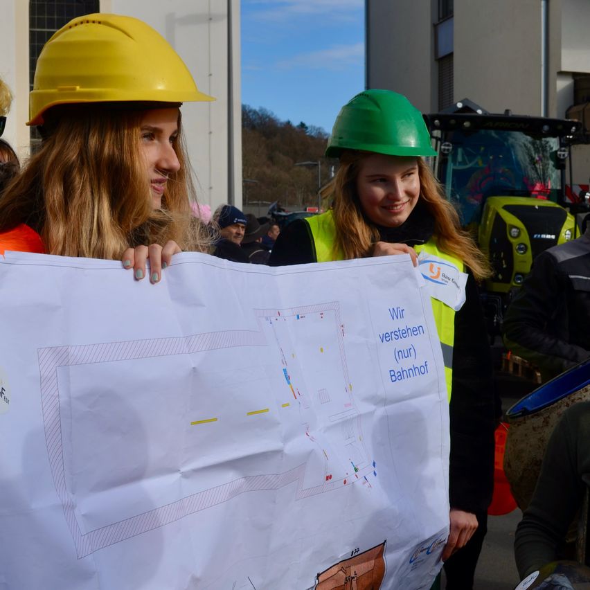 Zwei Frauen in Schutzhelmen halten ein großes weißes Plakat mit einem Diagramm und Text auf Deutsch. Sie lächeln und posieren für ein Foto vor einem Gebäude mit einem Traktor im Hintergrund.