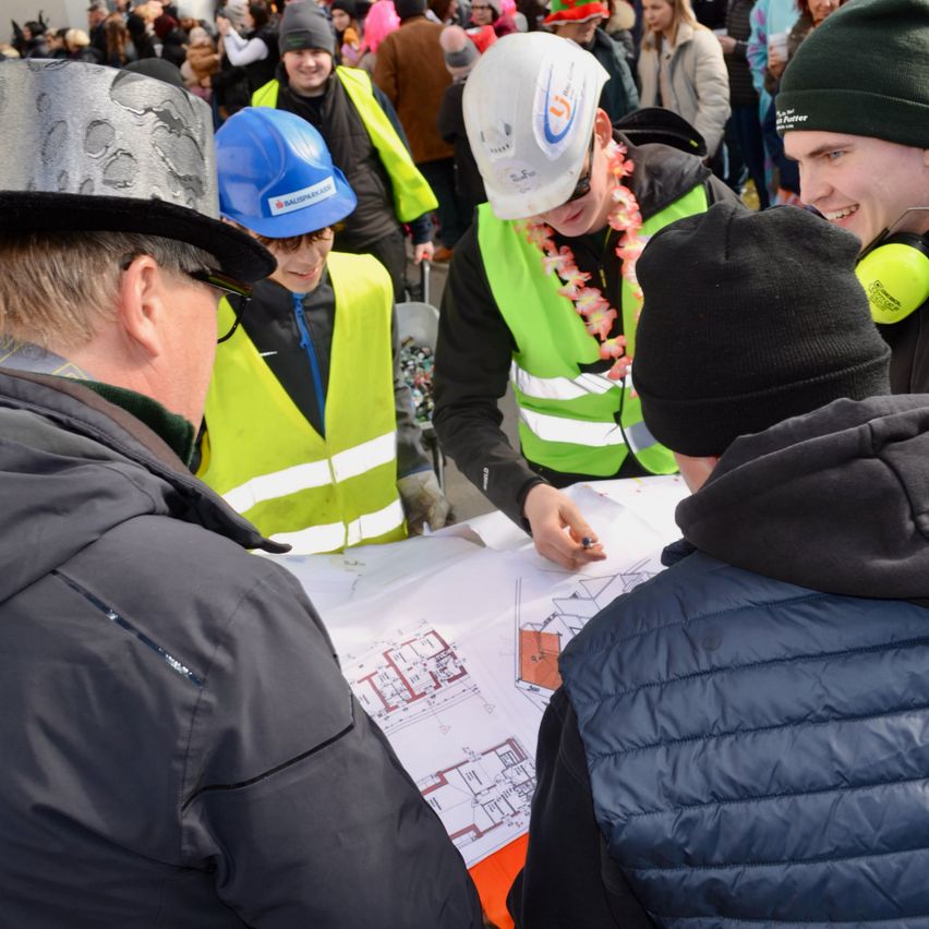 Mehrere Personen in Sicherheitswesten und Helmen, darunter eine mit einem weißen Helm, versammeln sich um ein großes Blatt Papier mit Architekturzeichnungen. Sie führen eine Diskussion.