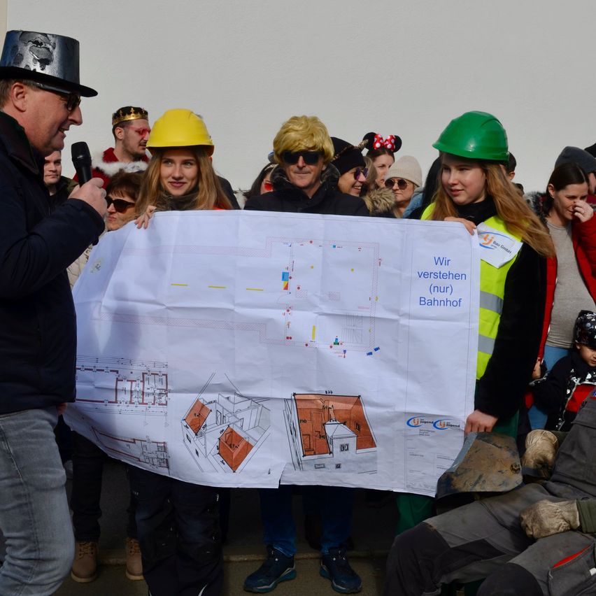 Ein Mann hält ein Mikrofon und spricht, während er neben einem großen Plakat steht, das von zwei Frauen in Bauhelmen gehalten wird. Sie sind von einer Menschenmenge umgeben, von denen einige festliche Kostüme tragen.