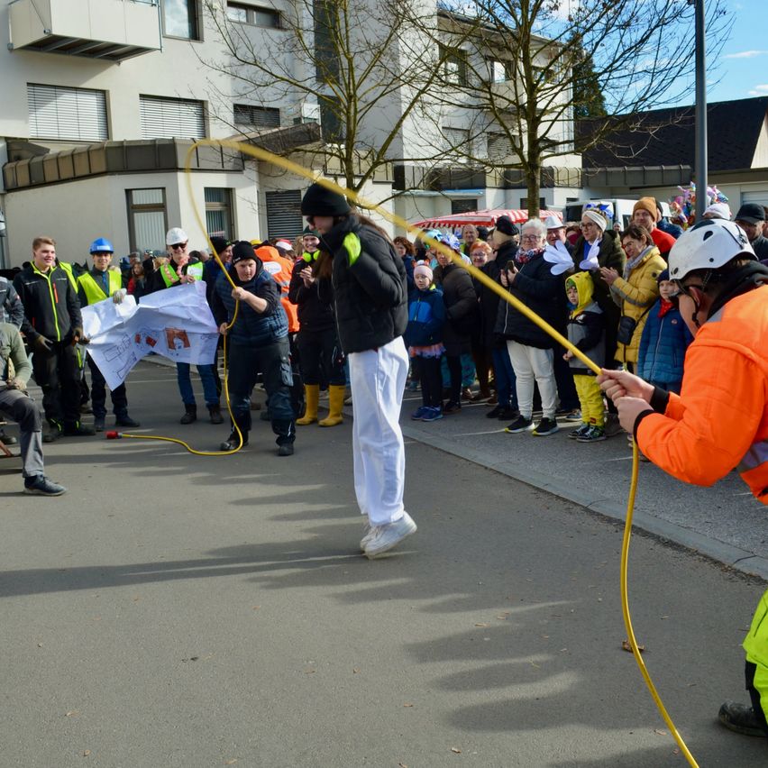 Eine Gruppe von Menschen auf einer Straße, einige in Sicherheitswesten, stehen um einen Mann mit einer Schnur. Der Mann zieht an der Schnur, und eine andere Person in einer orangefarbenen Weste hält das andere Ende. Im Hintergrund sind Gebäude zu sehen.