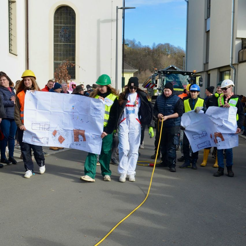 Eine Gruppe von Menschen in Arbeitskleidung mit Plänen und Bannern geht eine Straße hinunter. Ein Fahrzeug ist hinter ihnen geparkt.