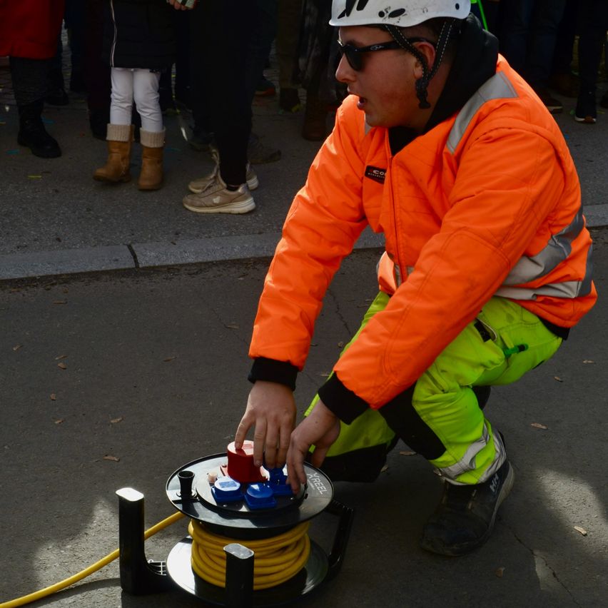 Ein Bauarbeiter in oranger und grüner Schutzkleidung kniet auf dem Boden und repariert möglicherweise etwas. Hinter ihm stehen Menschen in Winterkleidung.
