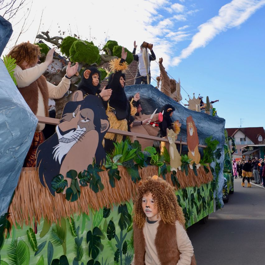Ein für einen Umzug geschmückter Wagen mit Menschen in Löwenkostümen, die winken, und einem Kind in Löwen-Make-up, das vorne geht.
