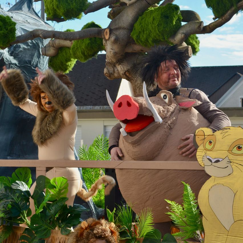 Eine Person in Löwenkostüm steht neben einer Person in Gnu-Kostüm und einer Löwen-Ausschnitt-Dekoration auf einem Umzugswagen.